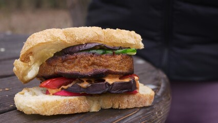 Delicious vegan burger made from plant-based meat substitute. Vegetables and a burger patty are grilled in the backyard. A juicy burger lies on a wooden table outside in the park.