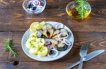 Cut smoked mackerel on plate with boiled potatoes, rings of onion, lemon, olives, oil, rosemary on wooden background