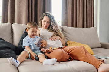 Young pregnant woman in casualwear sitting on soft comfortable couch next to her cute little son while both playing with toys