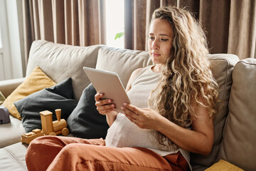 Young pregnant woman with digital tablet watching online video or blog for future mothers while...