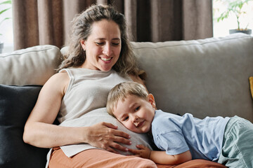 Fototapeta premium Happy adorable toddler boy listening to heartbeat of hitting of fetus in belly of his pregnant mother while keeping ear on her tummy