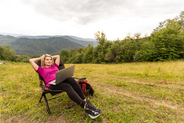 A woman in a sports warm suit works on a laptop outdoors in a mountainous area.