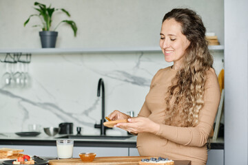 Fototapeta na wymiar Young smiling pregnant woman spreading honey or apple jam on slice of fresh wheat bread while preparing breakfast for herself