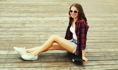 Portrait of happy smiling young woman with skateboard wearing shirt, sunglasses on city street background