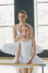 Young ballet teacher and her youthful learner in white tutu standing by wooden bar in front of mirror in classroom during repetition
