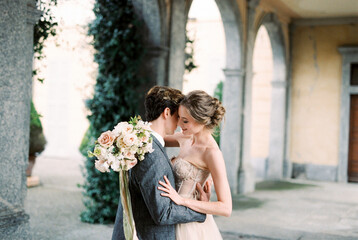 Groom hugs bride with a bouquet of flowers near the arches of an old villa. Como, Italy