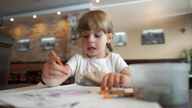 Little happy girl coloring a picture in a cafe. Everyday development of children. Leisure of the daughter while waiting.