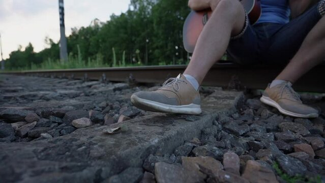 man musician in shorts sits on rail of railway plays on guitar taps his foot in beat of music outdoors