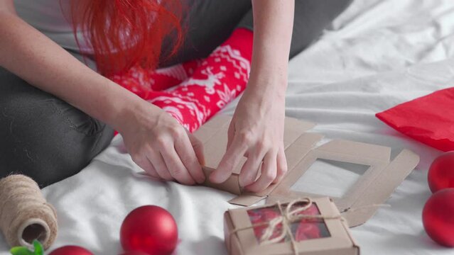 The girl makes a beautiful Christmas present on the bed. The best Christmas present ever. Women's hands make a gift box, prepare a surprise for the holiday. Red toy balls and New Year's hat.