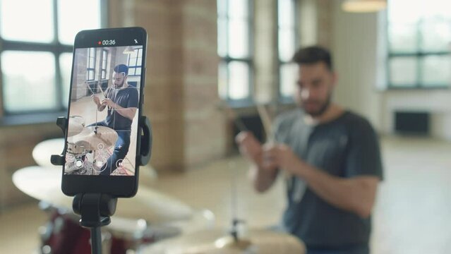 Rack focus shot of male drummer sitting at drum kit and speaking on camera while filming video lesson with smartphone on tripod