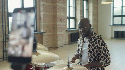 Rack focus shot of African American male drummer sitting at drum set and talking on camera while filming video lesson with smartphone on tripod