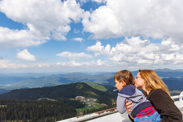 Fototapeta na wymiar Mother holds son and shows him landscapes of forest valley of Rhodope Mountains while standing on tower of Snezhan