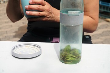 Wasserflasche mit grünen Minzeblättern auf weißem Tisch vor Oberkörper von Frau beim Essen bei Sonne am Mittag im Sommer
