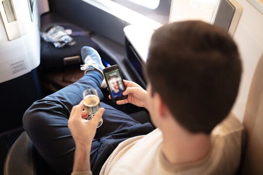 Rich man sitting on board of first class flight, drinking champagne and reading messages or checking business news on smartphone