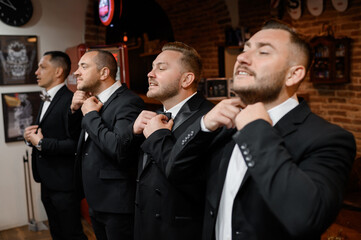 Side view of adult men and bridegroom in black suits, standing and together correcting their bow ties on necks in wedding morning