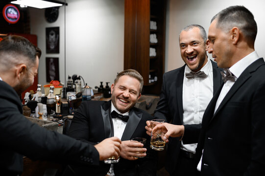 Front view of men and groom in suits, which having fun and clinking glasses with alcohol drinks while spending time together in barber shop during wedding morning