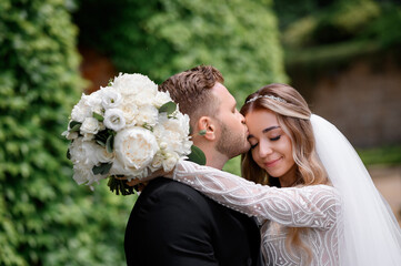 Close view of bearded groom in black suit, kissing bride which hugging his neck with flowers and closing eyes while posing on nature during wedding event