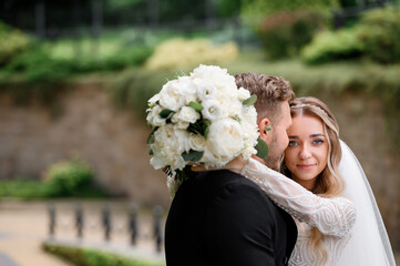 Close view of stunning woman with stylish hairdo and natural make up, embracing her husband and holding bridal bouquet on his back, smiling and looking at camera while posing on nature