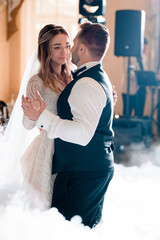 Side view of pensive bride with blonde curly hair, wearing in long lace dress, looking to her groom and dancing first wedding dance in hall among dry ice effect