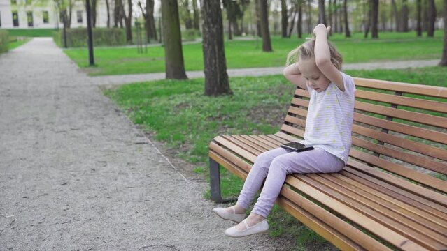 A girl sits on a park bench and ogles a video on her smartphone.