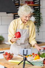 Senior woman cooking with video receipts or blogging. Mature female preparing fresh vitamin vegetarian salad with organic ingredients with a tutor in her smartphone. Elder using technology