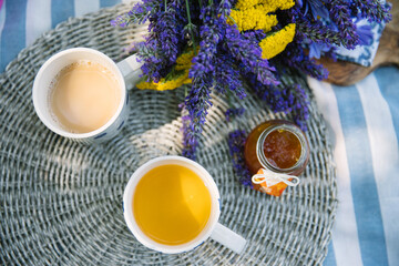 summer bouquet of yellow yarrow and violet lavender, 2 cups of tea with milk and jar of tangerine jam on a striped picnic blanket with summer vibe - 520679497
