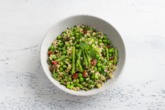 Traditional middle eastern or arabic tabbouleh salad of bulgur, tomatoes, asparagus, green peas and greens on light gray background, top view. Healthy homemade vegan food