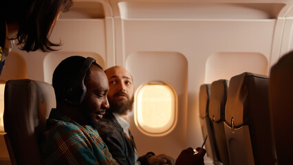 Male passenger talking to flight attendant on airplane, being seated on plane and using smartphone. Flying during sunset, travelling on vacation trip with tourists. © DC Studio