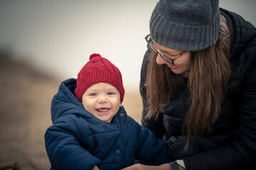Niño bebé con gorro de lana rojo y campera sonriendo y mirando a la cámara y jugando con su mamá en la playa en otoño e invierno