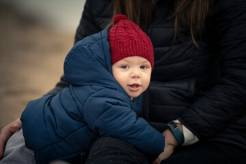 Niño bebé con gorro de lana rojo y campera mirando a la cámara y jugando con su mamá en la...