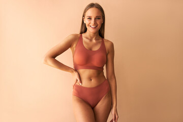 Attractive confident young woman posing on beige background in comfortable underwear and smiling. - obrazy, fototapety, plakaty