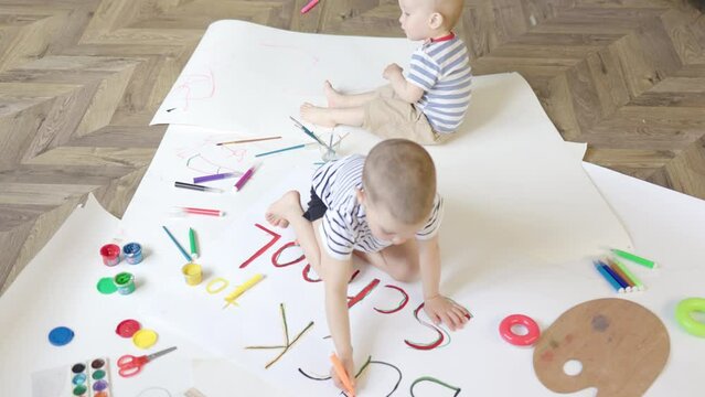 two sibling brothers boys sitting on floor indoors drawing arts, coloring words back to school. toddler baby boy helping brother do homework art class. family home education, distant learning kids