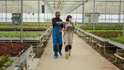 Agricultural engineer using laptop explaining to new african american worker the harvesting order for organic vegetables in greenhouse. Caucasian woman training employee on farming procedures.