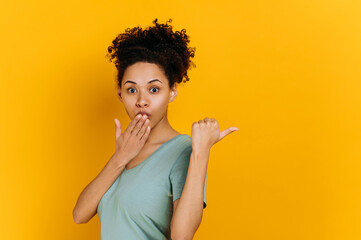 Shocked stunned african american curly haired young woman in a t-shirt, points finger to the side behind the back, at empty copy space, stands on isolated orange background, looks surprised at camera