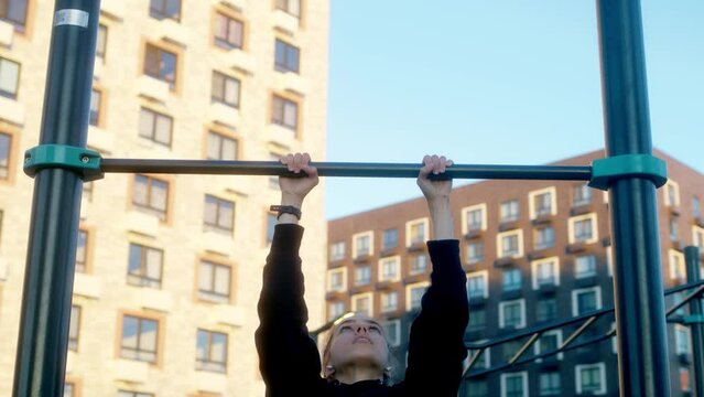 young woman in morning practices pull-ups on bar in black hoodie with pigtail. attractive brunette engaged in workout on sports ground in courtyard of house. close up view of hands pulling up