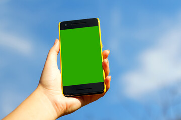 Woman hand holding black smartphone in yellow plastic case  on blue sky nature background, sunny day. Black screen. Mockup, mock-up, mock up. Female hand. Smart phone