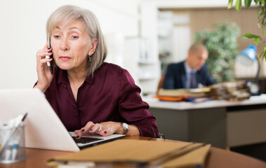 Concentrated elderly female entrepreneur solving business issues by phone, sitting in office with laptop..