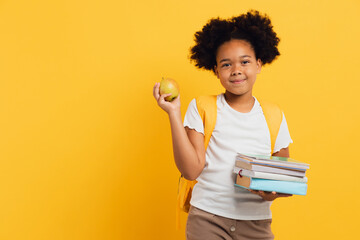 Happy African american schoolgirl holding notebooks, books and lunch apple on yellow background, copy space.
