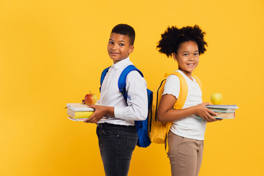 Happy african american schoolgirl and mixed race schoolboy holding books side by side on yellow background. Back to school concept.
