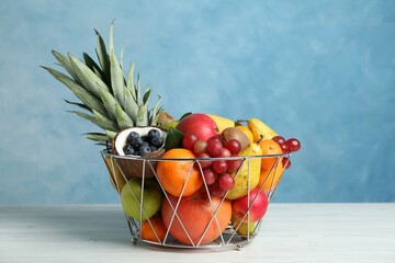 Assortment of fresh exotic fruits in metal basket on white wooden table against light blue...