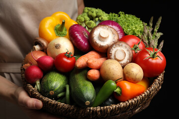 Woman holding wicker basket full of fresh vegetables on black background, closeup