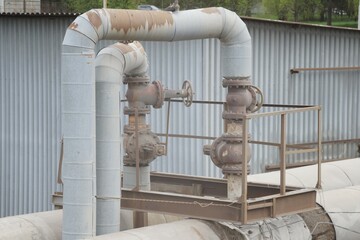 Full-color horizontal photo. Tie-in to the main pipeline. Thin pipes extending from the main pipes. 4 rusty Gate valves and a platform for staff are visible.