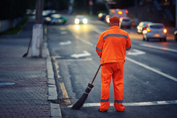 Man with broom clean city road and roadside at night. Worker cleans roads, streets, footpaths with...