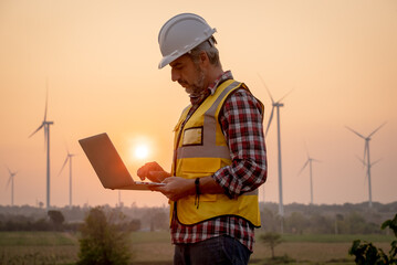 Fototapeta Portrait of engineer wearing yellow vest and white helmet using a computer laptop on site at wind turbines field or farm, Sustainable energy concept obraz