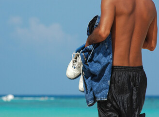 Rear view of a young Mexican adult observing the sea from the beach, clothing in his hands. Summer vacation concept