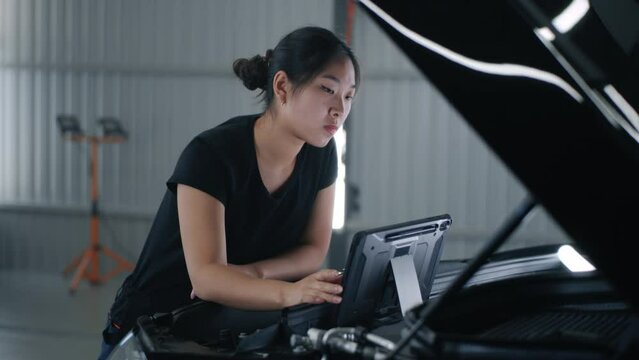 Asian female mechanic in uniform checking car engine and swiping on tablet computer during diagnostics and maintenance in car service