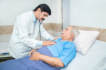 Indian doctor wearing stethoscope check heart beat of olde senior male patient lying on bed at...