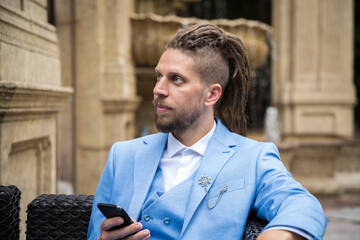 Young successful hipster caucasian man with dreadlocks in a blue suit with a smartphone in his hands sits in a street restaurant looks away. - Powered by Adobe