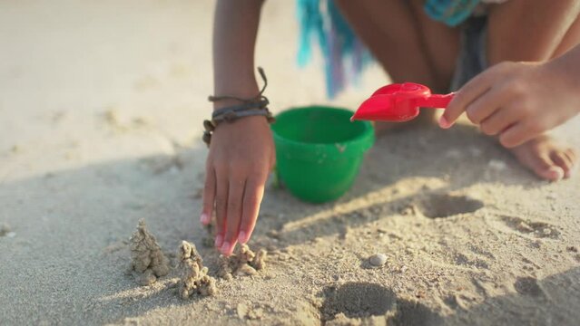 A girl with african braids in a summer costume plays on the beach with shells near the sea with waves under sunny sunset