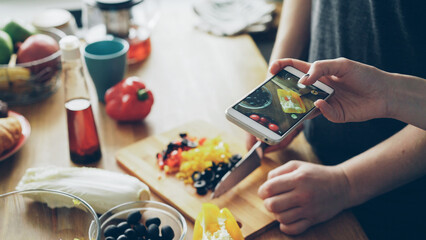 Fototapeta na wymiar Closeup of male hands cutting vegetables for healty breakfast on table and his girlfriend taking photos with smartphone camera for social media